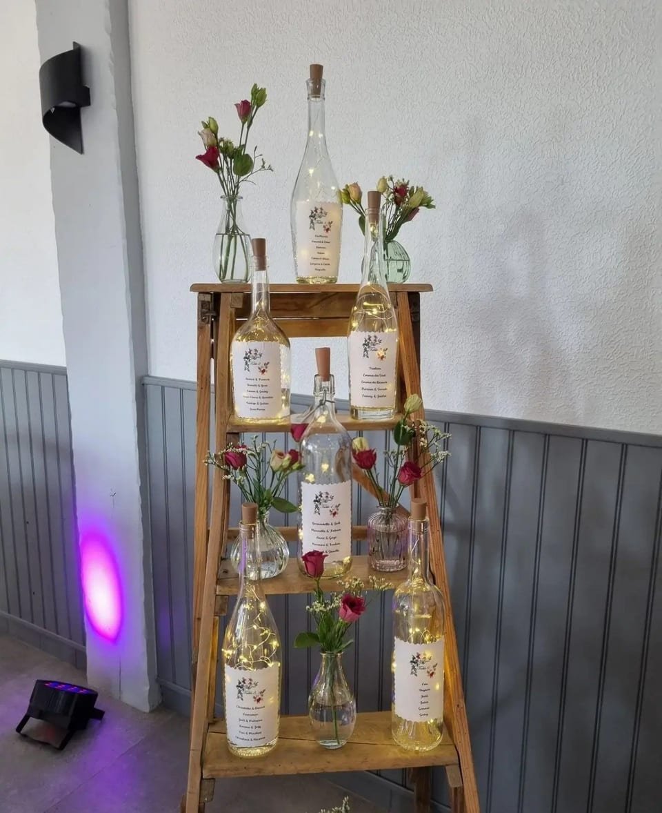Plan de table présenté sur un escabeau ancien, bouteilles lumineuses et fleurs